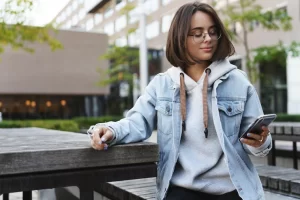 portrait-young-modern-hipster-girl-denim-jacket-glasses-sitting-outdoors-city-square-park-bench-turn-aside-check-messages-mobile-phone-smiling-as-texting-friend-use-application_197531-30562